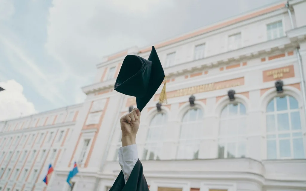 A hand holding a black graduation cap aloft before a large university building